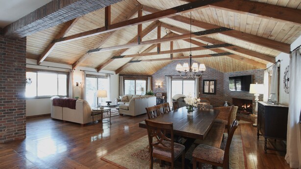 A spacious living room with a high wooden ceiling, a fireplace, and a dining area with a long table and chairs.
