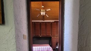 Kapea käytävä johtaa makuuhuoneeseen, jossa on sänky, kattotuuletin ja puupanelointi.