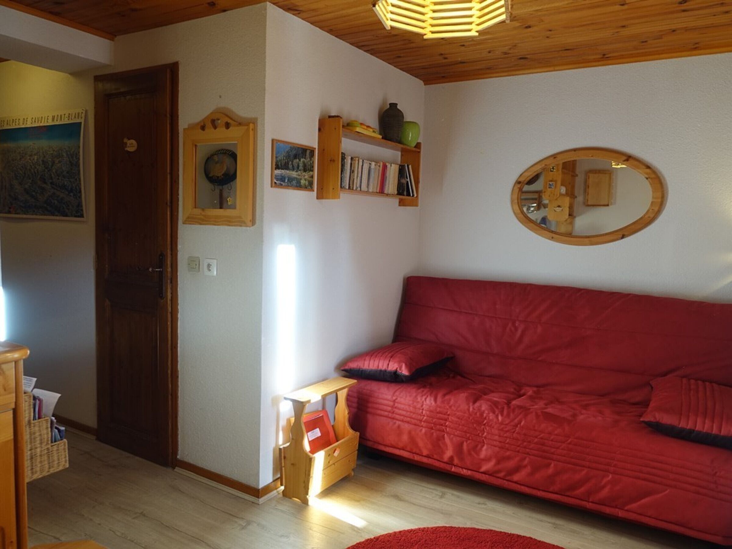 Un salon chaleureux avec un canapé rouge, des étagères en bois ornées de livres et d’objets décoratifs, une petite table en bois et un miroir accroché au mur.