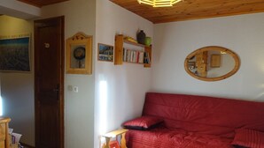 A cozy living room with a red sofa, wooden shelves with books and decorative items, a wooden side table, and a mirror on the wall. - Charming 4-5 Person Apartment, Well-Equipped, Central Location, Elevator, 50m to Slopes & Shops (Arêches-Beaufort)