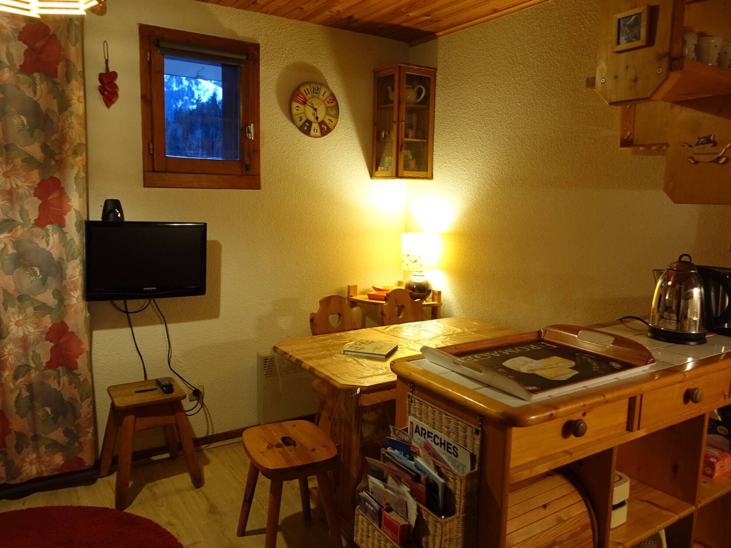 A cozy kitchen with a wooden table, chairs, a television, a clock, and a window with a view of trees.