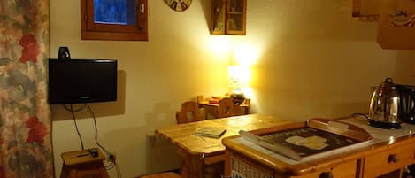 A cozy kitchen with a wooden table, chairs, a television, a clock, and a window with a view of trees.