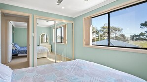 A bedroom with a large window, a bed with patterned bedding, a mirror, and a view of a building and trees outside. - 3-45 Park Street - Spacious Townhouse (EVANS HEAD)