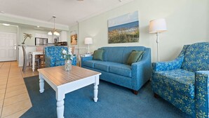 Una sala de estar con un sofá azul, un sillón estampado, una mesa de centro de madera y un cuadro de una playa.