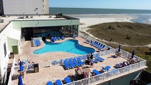 Una piscina en la azotea con sillas reclinables azules, un bar con paredes de cristal y vista a la playa.