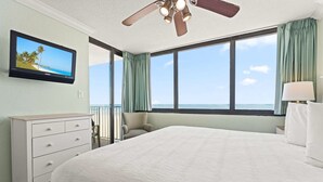 Habitación de hotel con cama, televisión, ventilador de techo, ventana con cortinas y vista al océano.