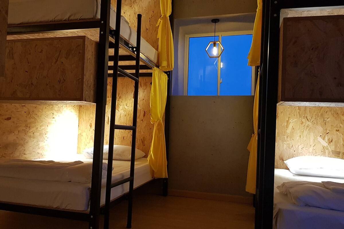 A dormitory room with bunk beds, a window with yellow curtains, and a hanging light fixture.