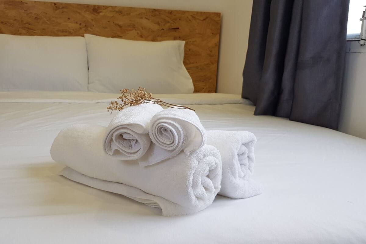 A neatly made bed with white linens and rolled towels, a wooden headboard, and a dark curtain.