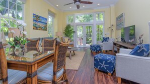 A spacious living room with a glass dining table, wicker chairs, a ceiling fan, and a large window with a view of palm trees. - Island Oasis at the Truman Annex in Key West (Key West)