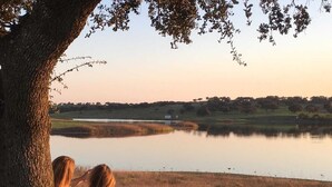 Un banco con dos personas sentadas, con vista a un lago tranquilo, árboles y una colina distante.