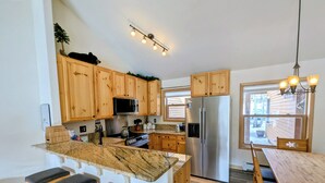 Una cocina con armarios de madera, un refrigerador de acero inoxidable, una encimera de mármol y un comedor con mesa y sillas de madera.