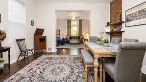 A spacious living room with a wooden dining table, grey upholstered chairs, a patterned rug, and a brick accent wall. - Centrally located in the Tower Grove East neighborhood! (St. Louis)