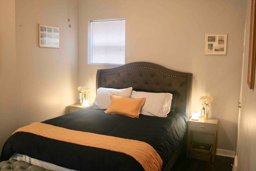 A bedroom with a large bed, two bedside tables with lamps, a window with blinds, and framed pictures on the walls.