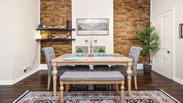 A dining area with a wooden table, grey upholstered chairs, a bench, a patterned rug, a brick wall, a framed picture, and a potted plant. - Centrally located in the Tower Grove East neighborhood! (St. Louis)