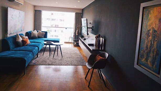Een moderne woonkamer met een blauwe hoekbank, een klein rond tafeltje en een televisie aan een donkere muur.