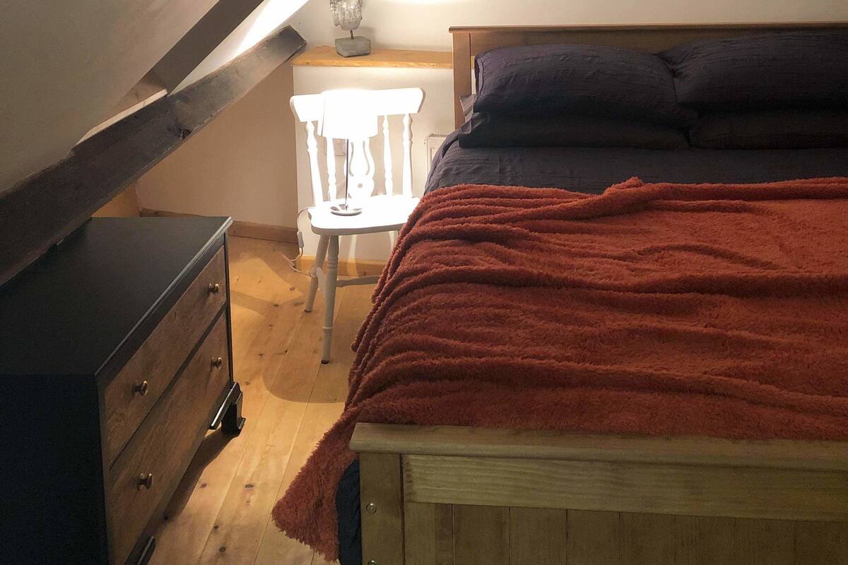Un dormitorio con techo inclinado, una cama con manta roja, un aparador de madera y una silla blanca.