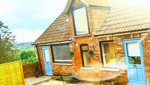 A two-story brick house with blue doors and windows, a sloped roof, and a garden area with potted plants and a wheelbarrow. - The Horse Stables (Glastonbury)