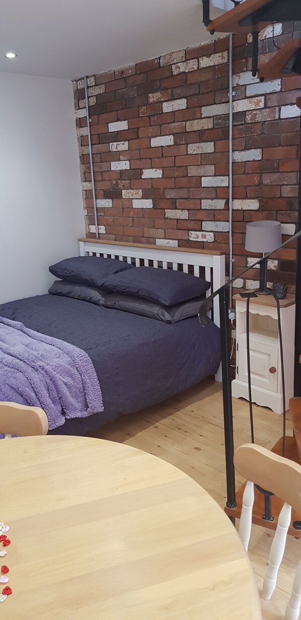 A bedroom with a brick-patterned wall, a bed with a white headboard, a bedside table with a lamp, and a small wooden table with chairs. - The Horse Stables (Glastonbury)