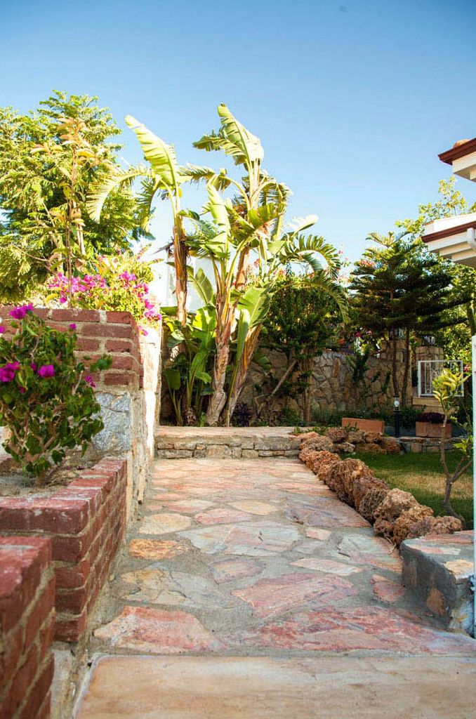 Un chemin de pierres mène à une maison avec un mur en briques et une végétation luxuriante, notamment des bananiers et des plantes à fleurs.