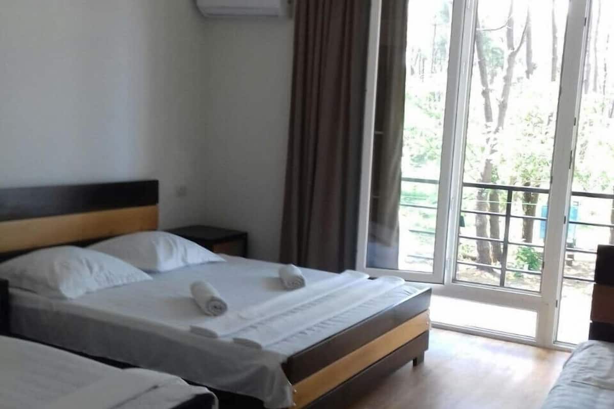 Quarto de hotel com duas camas, piso de madeira, janela com cortinas e um ar condicionado na parede.