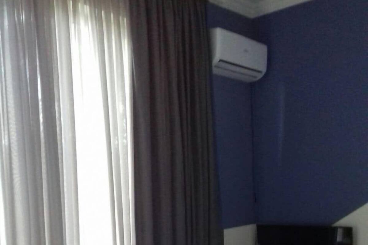 Uma cama de solteiro com colchão e travesseiro brancos, uma mesinha de cabeceira, uma janela com cortinas e um ar condicionado na parede.