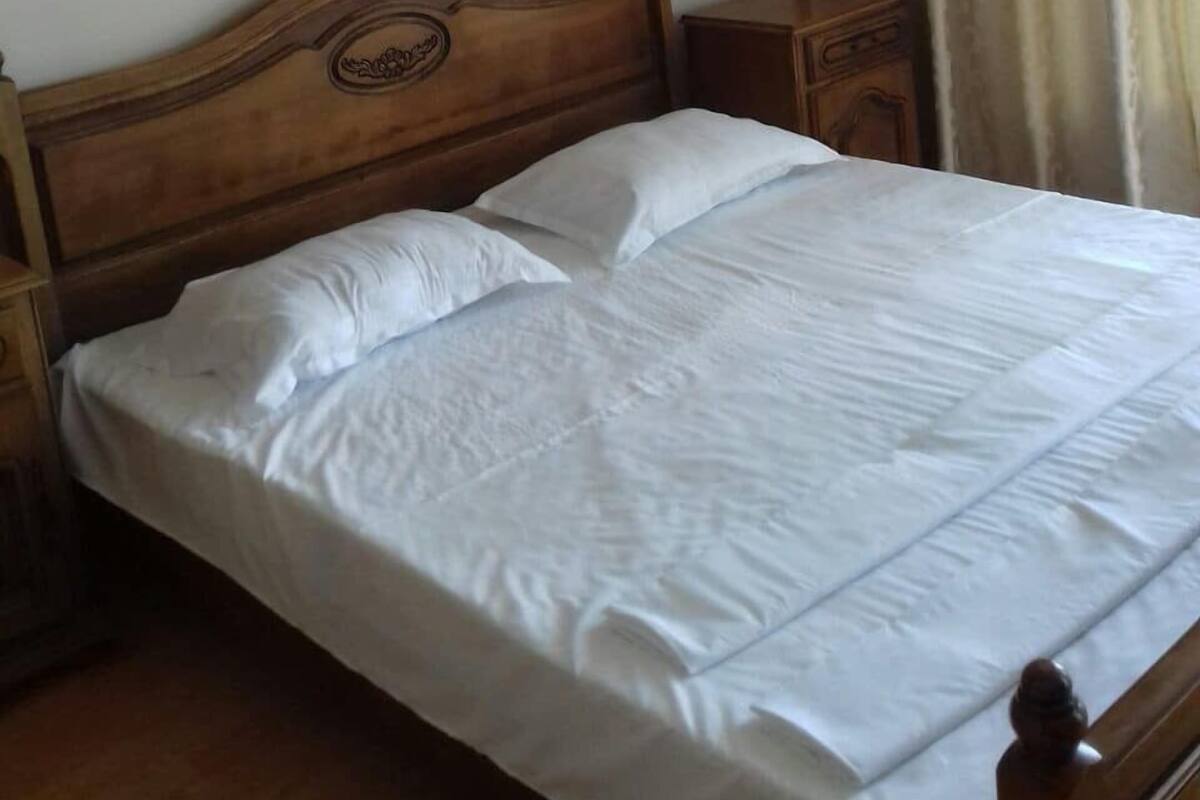 Uma cama bem arrumada com cabeceira de madeira e dois mesinhas de cabeceira, uma com gaveta e outra com uma pequena mesa.