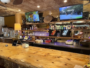 Una zona de bar con mostrador de madera, varias pantallas y una variedad de botellas y vasos.