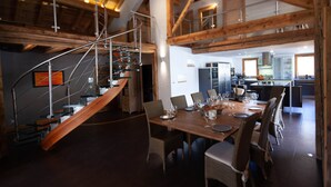 Une salle à manger avec une table en bois dressée pour six personnes, un escalier avec des rampes en verre et, au fond, une cuisine.