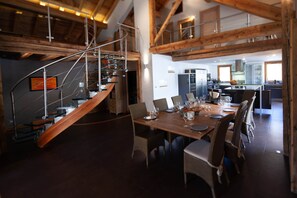 A dining area with a wooden table set for six, a staircase with glass railings, and a kitchen in the background.
