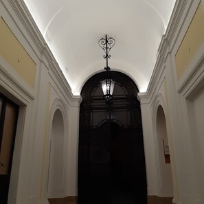 Un pasillo con techo alto, un candelabro decorativo y un vano oscuro al final.
