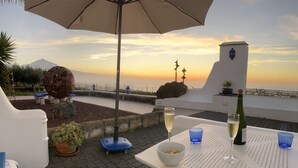 Une terrasse avec une table dressée pour deux personnes, agrémentée d’une bouteille de vin et de verres, offrant une vue panoramique sur un paysage magnifique, avec un coucher de soleil et une montagne lointaine.
