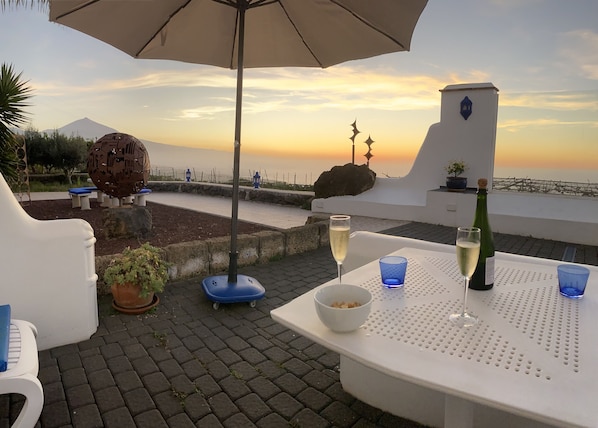 A terrace with a table set for two, featuring a bottle of wine and glasses, overlooking a scenic view with a sunset and a distant mountain.