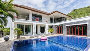Una casa moderna de dos pisos con piscina, mobiliario exterior y montañas al fondo.