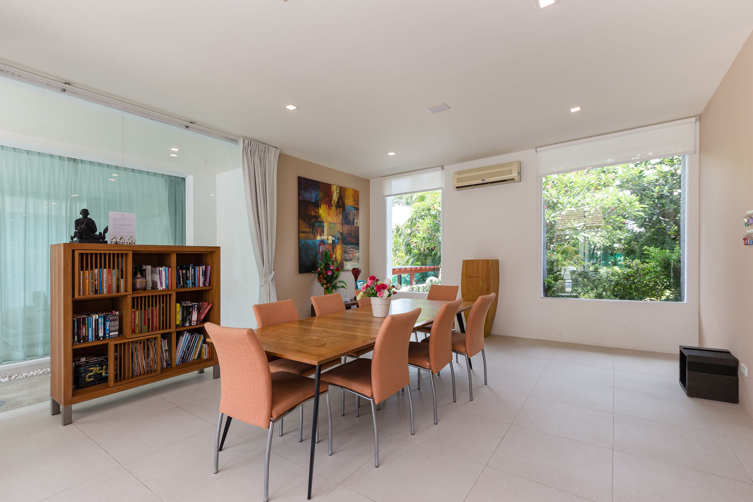 A modern dining area with a wooden table and chairs, a bookshelf, and large windows with a view of greenery.