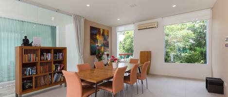 A modern dining area with a wooden table and chairs, a bookshelf, and large windows with a view of greenery.