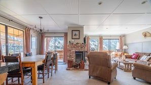 A spacious living room with a fireplace, a dining area with a wooden table and chairs, and large windows offering a view of the mountains.