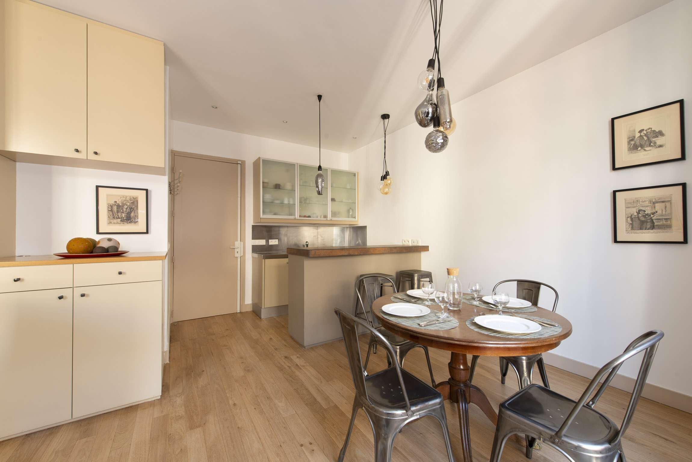 A dining area with a round wooden table, metal chairs, and a kitchen in the background.