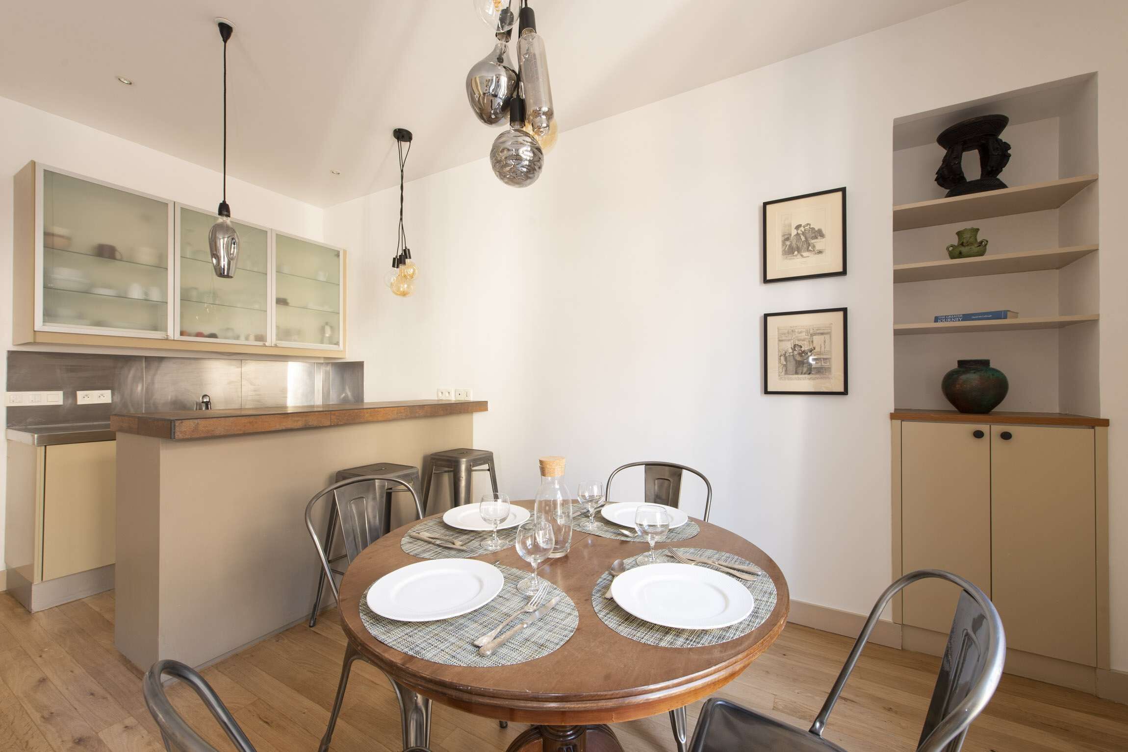 Un comedor con una mesa redonda dispuesta para cuatro personas, rodeada de sillas metálicas. Se ve una cocina al fondo con una encimera de madera y un armario de vidrio.