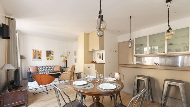Una sala moderna con un comedor listo para cuatro personas, rodeado de sillas de metal. Hay un sofá, una mesa de centro y un televisor en la sala. Se ve la cocina, con electrodomésticos de acero inoxidable y una ventana sobre el fregadero.