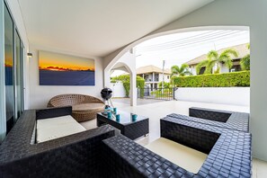 A modern outdoor seating area with black cushioned sofas and a wicker chair, a glass coffee table, and a painting of a sunset. - Modern 4 Bedrooms Pool Villa - VY (Hua Hin)