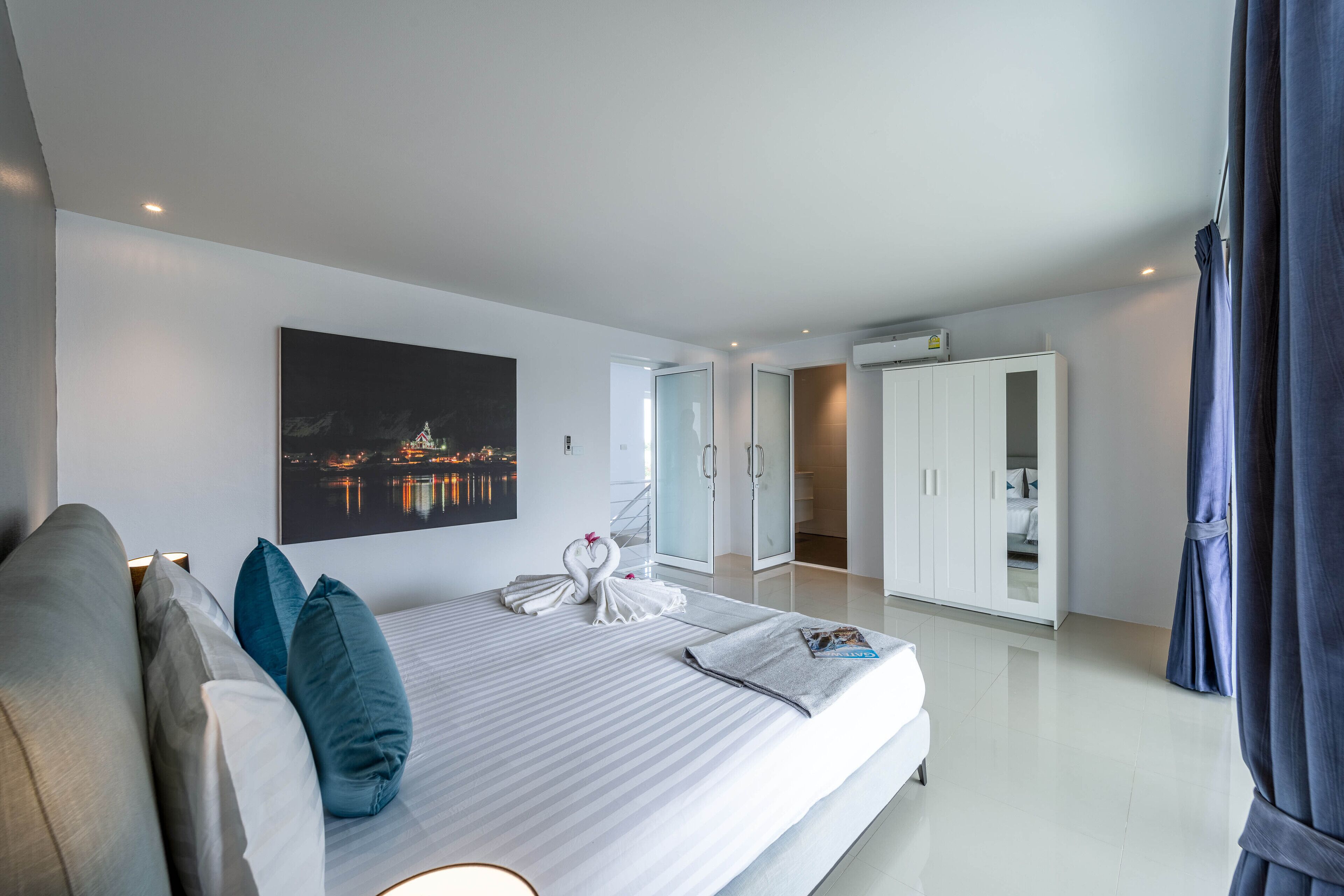 A modern hotel room with a large bed, a night view painting, a wardrobe, and a bathroom visible through an open door.