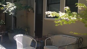 En gårdsplads med to metalstole og et bord, omgivet af krukker med planter og en bygning med store vinduer.