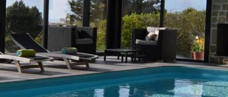 En modern inomhuspool med klart vatten, stora fönster och solstolar.