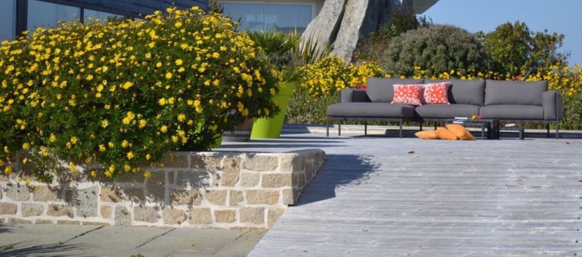 A patio area with a stone planter, a gray sofa with red and white pillows, and yellow flowers.