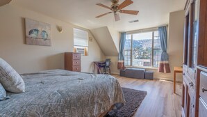 Un dormitorio con cama, escritorio, silla, ventana con cortinas, ventilador de techo y vistas al exterior.