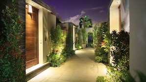 Un sendero bien iluminado bordeado por abundante vegetación y arquitectura moderna, que conduce a una puerta de madera.