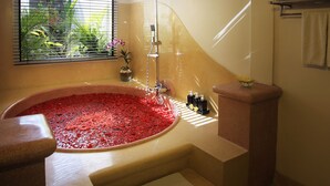 Un baño con una bañera grande llena de guijarros rojos, un cabezal de ducha y una ventana con persianas.