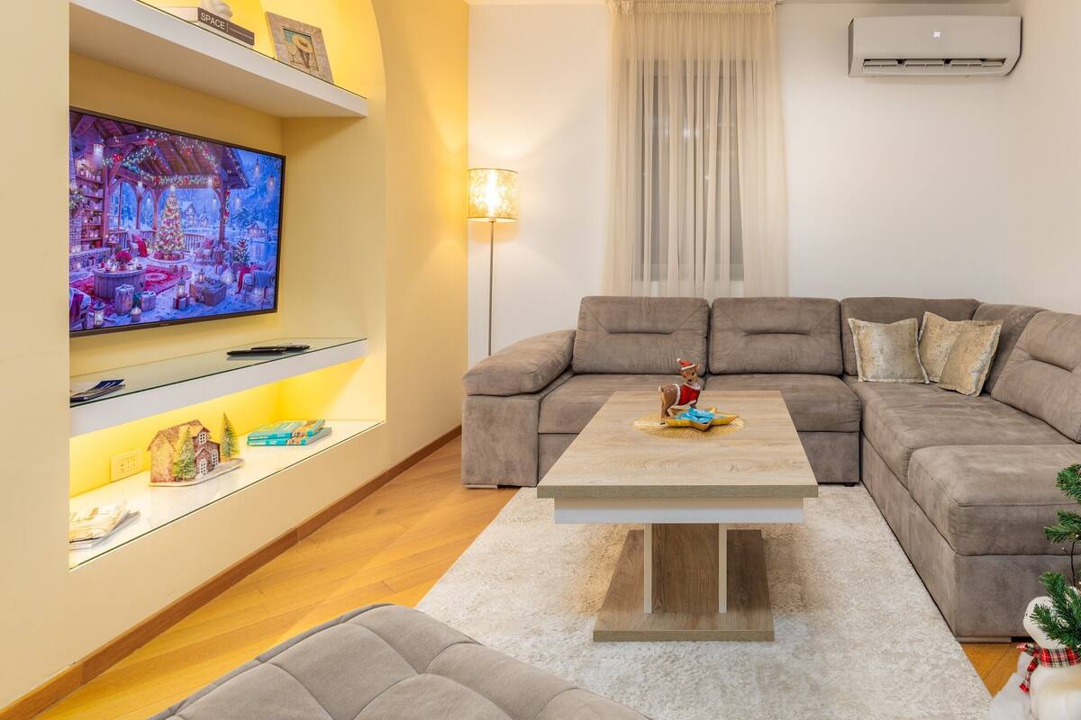 Ein modernes Wohnzimmer mit einem Ecksofa, einem Flachbildfernseher und einer festlichen Weihnachtsdekoration an der Wand.