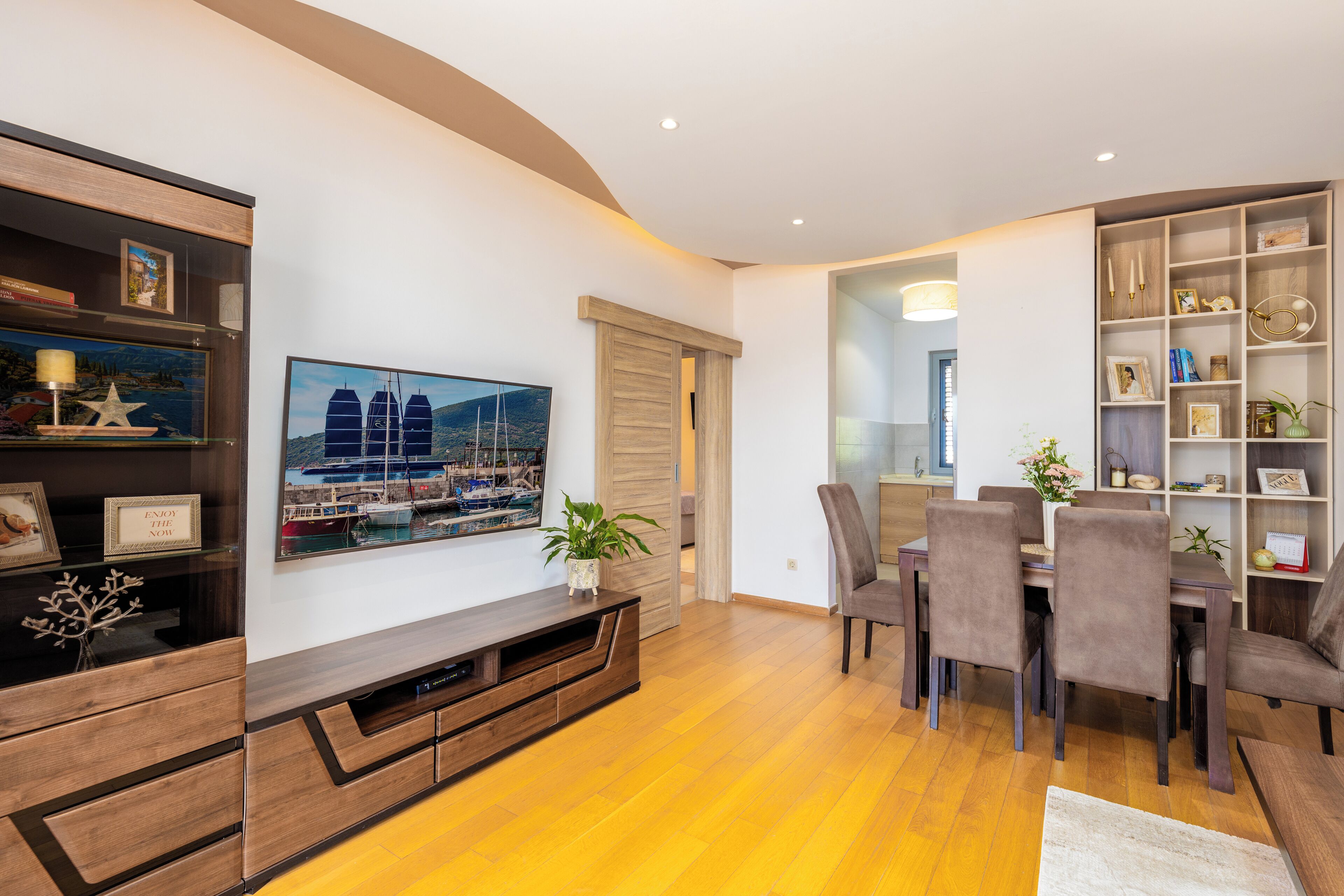 Een moderne woonkamer met een houten vloer, een eethoek met een tafel en stoelen, en een boekenkast met diverse spullen.