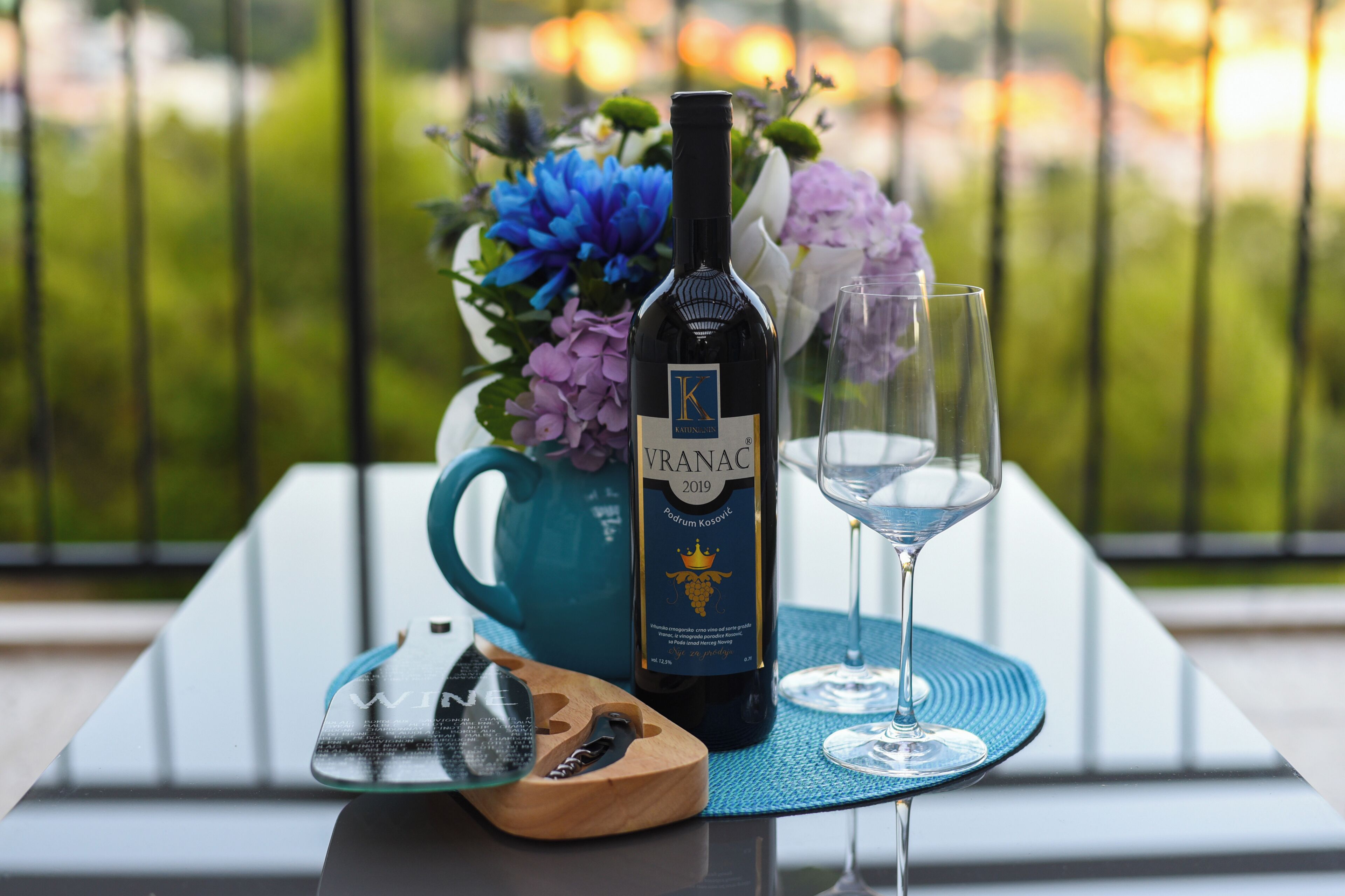 Una botella de vino Vranac, una copa y un sacacorchos de madera sobre una mesa con una servilleta azul, frente a un balcón con vistas.
